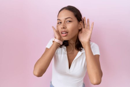 Téléchargez les photos : Jeune femme hispanique portant un t-shirt blanc décontracté essayant d'entendre les deux mains sur le geste de l'oreille, curieuse de commérages. problème auditif, sourd - en image libre de droit