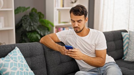 Foto de Hombre hispano joven usando teléfono inteligente sentado en el sofá en casa - Imagen libre de derechos