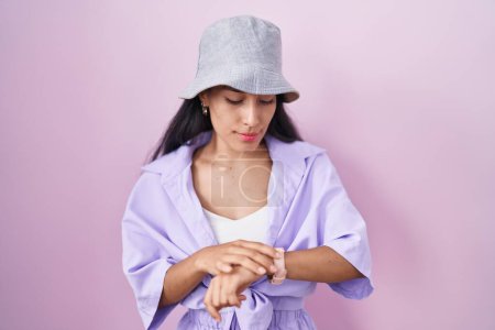 Foto de Mujer hispana joven de pie sobre fondo rosa con sombrero que comprueba la hora en el reloj de pulsera, relajado y seguro - Imagen libre de derechos