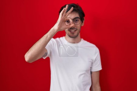 Téléchargez les photos : Jeune homme hispanique debout sur fond rouge faisant un geste correct avec la main souriante, les yeux regardant à travers les doigts avec un visage heureux. - en image libre de droit