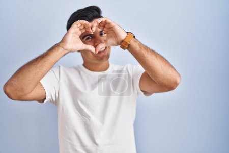 Photo for Hispanic man standing over blue background doing heart shape with hand and fingers smiling looking through sign - Royalty Free Image