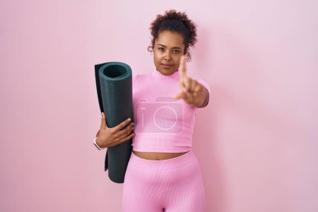 Foto de Mujer hispana joven con el pelo rizado sosteniendo estera de yoga sobre fondo rosa señalando con el dedo hacia arriba y la expresión enojada, mostrando ningún gesto - Imagen libre de derechos