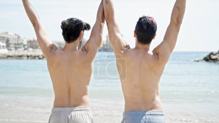Téléchargez les photos : Deux hommes couple touristique debout vers l'arrière avec les mains levées à la plage - en image libre de droit