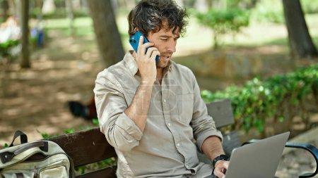 Photo for Young hispanic man talking on smartphone using laptop sitting on bench at park - Royalty Free Image