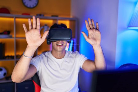 Téléchargez les photos : Jeune homme hispanique streamer jouer à un jeu vidéo en utilisant des lunettes de réalité virtuelle à la salle de jeu - en image libre de droit