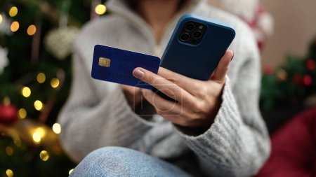 Foto de Joven mujer hispana hermosa usando teléfono inteligente y tarjeta de crédito sentado en el sofá por el árbol de Navidad en casa - Imagen libre de derechos