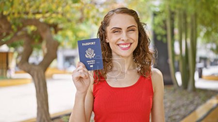 Foto de Joven confiada sonriendo alegremente, orgullosamente sosteniendo su pasaporte americano en el parque verde, lista para un viaje de vacaciones soleado. - Imagen libre de derechos