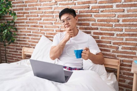 Foto de Joven asiático hombre beber café sentado en la cama seria cara pensando en la pregunta con la mano en la barbilla, reflexivo acerca de la idea confusa - Imagen libre de derechos