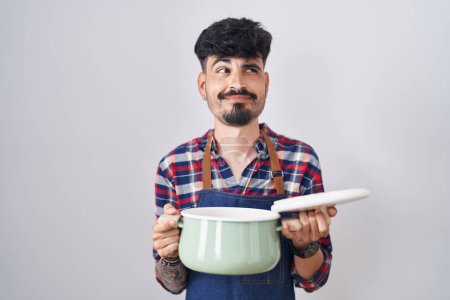Téléchargez les photos : Jeune homme hispanique avec la barbe portant tablier tenant casserole souriant regardant vers le côté et regardant loin en pensant. - en image libre de droit