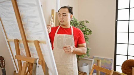 Foto de Hombre chino joven guapo, artista seguro de sí mismo, dibujo en el interior, inmerso en la creatividad en el estudio de arte con pincel en la mano, adornado con peinado tradicional de la coleta y delantal - Imagen libre de derechos