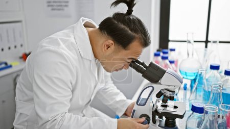 Téléchargez les photos : Un jeune Chinois sérieux, un scientifique professionnel, se concentrant au microscope dans un laboratoire animé - un mariage de médecine et de technologie - en image libre de droit