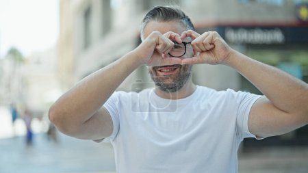 Téléchargez les photos : Homme aux cheveux gris souriant confiant faire geste de cœur avec les mains à la rue - en image libre de droit