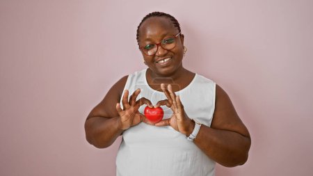 Téléchargez les photos : Femme afro-américaine confiante tient joyeusement la forme du cœur avec les mains, souriant sur fond de mur rose isolé, exprimant un mode de vie positif - en image libre de droit