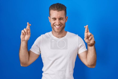 Téléchargez les photos : Jeune homme caucasien debout sur fond bleu geste doigt croisé souriant avec espoir et les yeux fermés. chance et concept superstitieux. - en image libre de droit