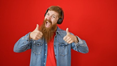 Téléchargez les photos : Jeune rousse homme écoutant de la musique faisant pouce vers le haut geste sur fond rouge isolé - en image libre de droit