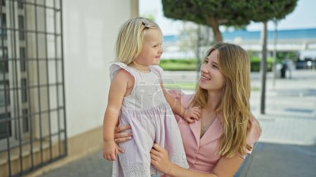 Photo for Cheerful caucasian mother and daughter embracing a warm, loving hug, sitting on a city bench, laughing, and smiling together, filled with joy and happiness, outdoors on a sunny street. - Royalty Free Image