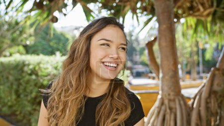 Téléchargez les photos : Jolie jeune femme hispanique, pleine de confiance, rayonnante d'un sourire radieux, debout à l'extérieur dans le parc verdoyant en s'amusant, les yeux joyeusement tournés vers le côté - en image libre de droit