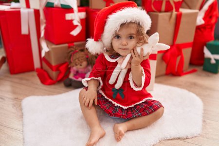 Téléchargez les photos : Adorable tout-petit blonde tenant une poupée de lapin assise sur le sol par des cadeaux de Noël à la maison - en image libre de droit