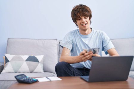 Téléchargez les photos : Jeune homme blond utilisant un ordinateur portable tenant des dollars à la maison - en image libre de droit