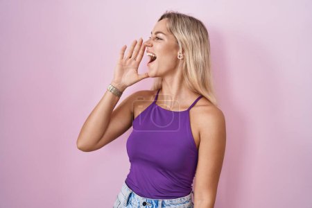 Photo for Young blonde woman standing over pink background shouting and screaming loud to side with hand on mouth. communication concept. - Royalty Free Image