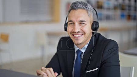 Téléchargez les photos : Jeune homme hispanique travailleur d'affaires à l'aide d'écouteurs souriant au bureau - en image libre de droit