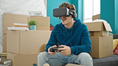 Téléchargez les photos : Jeune homme hispanique jouer à un jeu vidéo en utilisant joystick et lunettes de réalité virtuelle à la nouvelle maison - en image libre de droit