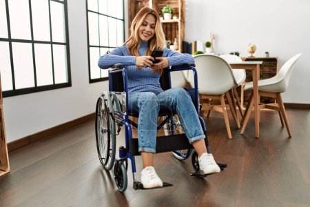 Téléchargez les photos : Jeune femme caucasienne utilisant smartphone assis sur fauteuil roulant à la maison - en image libre de droit