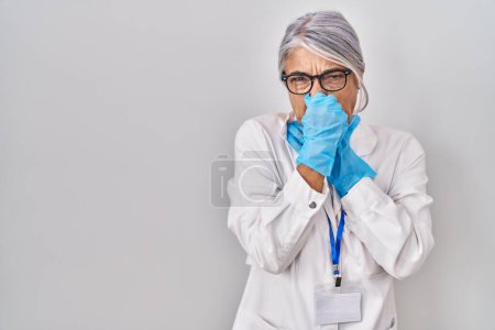 Téléchargez les photos : Femme d'âge moyen aux cheveux gris portant une robe de scientifique sentant quelque chose de puant et dégoûtant, odeur intolérable, retenant son souffle avec les doigts sur le nez. mauvaise odeur - en image libre de droit