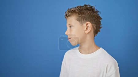 Photo for Adorable little blond boy, a cute caucasian kid, looking to the side with a serious expression on his face. standing, relaxed yet concentrated, isolated against a blue background wall. - Royalty Free Image