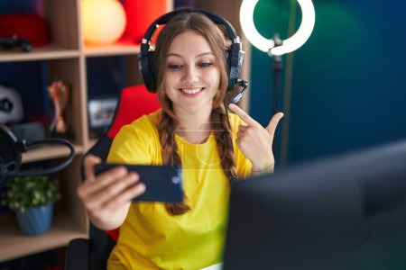 Foto de Joven mujer caucásica jugando videojuegos con smartphone sonriendo feliz apuntando con la mano y el dedo - Imagen libre de derechos