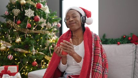 Foto de Mujer afroamericana escuchando música tomando café sentado junto al árbol de Navidad en casa - Imagen libre de derechos