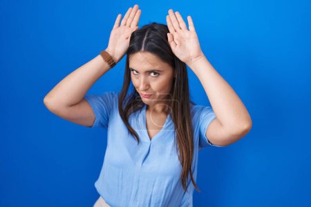 Photo for Young brunette woman standing over blue background doing bunny ears gesture with hands palms looking cynical and skeptical. easter rabbit concept. - Royalty Free Image