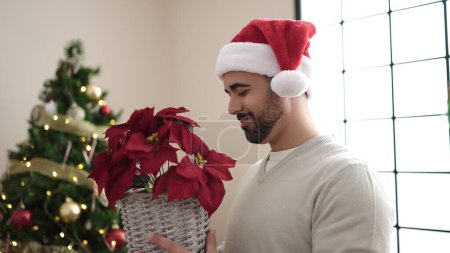 Téléchargez les photos : Jeune homme hispanique tenant une plante debout près de l'arbre de Noël à la maison - en image libre de droit
