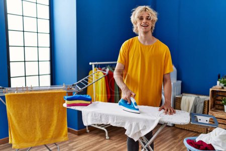 Photo for Young blond man smiling confident ironing clothes at laundry room - Royalty Free Image