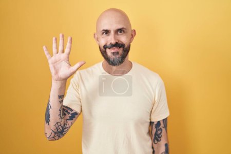 Téléchargez les photos : Homme hispanique avec des tatouages debout sur fond jaune montrant et pointant vers le haut avec les doigts numéro cinq tout en souriant confiant et heureux. - en image libre de droit