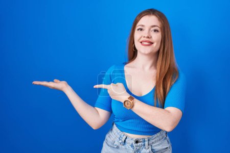 Téléchargez les photos : Femme rousse debout sur fond bleu étonné et souriant à la caméra tout en présentant avec la main et pointant du doigt. - en image libre de droit