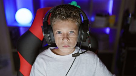 Photo for Adorable blond boy streamer, serious face focused on game, playing in dark gaming room with digital tech, sporting headset and sitting at computer table - Royalty Free Image
