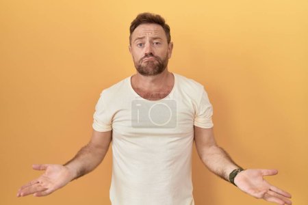 Photo for Middle age man with beard standing over yellow background clueless and confused with open arms, no idea concept. - Royalty Free Image