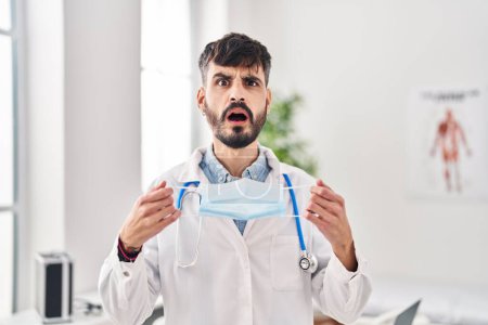 Foto de Joven médico hispano con barba sosteniendo máscara de seguridad en la cara de choque, mirando escéptico y sarcástico, sorprendido con la boca abierta - Imagen libre de derechos