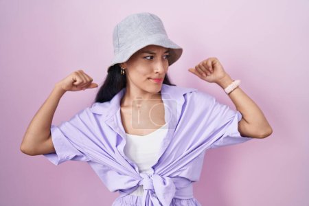 Photo for Young hispanic woman standing over pink background wearing hat showing arms muscles smiling proud. fitness concept. - Royalty Free Image