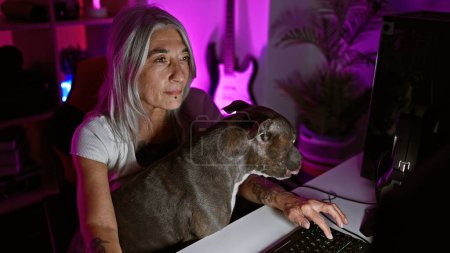 Téléchargez les photos : Focus féroce, femme d'âge moyen aux cheveux gris streamer jouer sérieusement à des jeux sur le ruisseau, chien à ses côtés dans une salle de jeux confortable - en image libre de droit