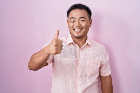 Foto de Joven chino de pie sobre fondo rosa haciendo el gesto feliz pulgares hacia arriba con la mano. aprobando la expresión mirando a la cámara mostrando éxito. - Imagen libre de derechos