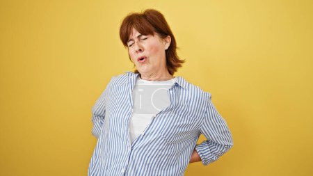 Téléchargez les photos : Femme d'âge moyen souffrant de maux de dos debout sur fond jaune isolé - en image libre de droit