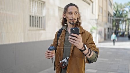 Téléchargez les photos : Jeune homme hispanique touriste en utilisant smartphone tenant café à la rue - en image libre de droit