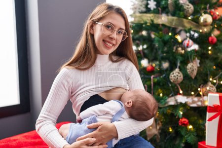 Photo for Mother and son breastfeeding baby standing by christmas tree at home - Royalty Free Image