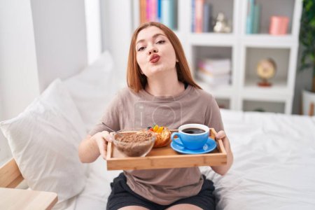 Téléchargez les photos : Femme rousse en pyjama tenant le plateau de petit déjeuner en regardant la caméra souffler un baiser étant belle et sexy. expression d'amour. - en image libre de droit