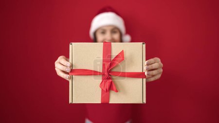 Téléchargez les photos : Jeune belle femme hispanique portant un chapeau de Noël tenant cadeau sur fond rouge isolé - en image libre de droit