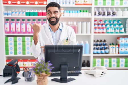 Téléchargez les photos : Homme hispanique avec barbe travaillant à la pharmacie pharmacie souriant avec le visage heureux regardant et pointant vers le côté avec le pouce vers le haut. - en image libre de droit