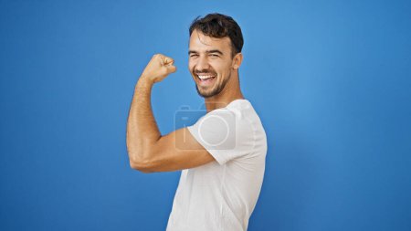 Téléchargez les photos : Jeune homme hispanique souriant confiant faire un geste fort avec le bras sur fond bleu isolé - en image libre de droit