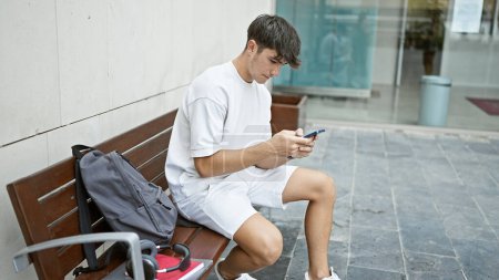 Téléchargez les photos : Jeune adolescent hispanique étudiant utilisant un smartphone assis sur un banc à l'université - en image libre de droit